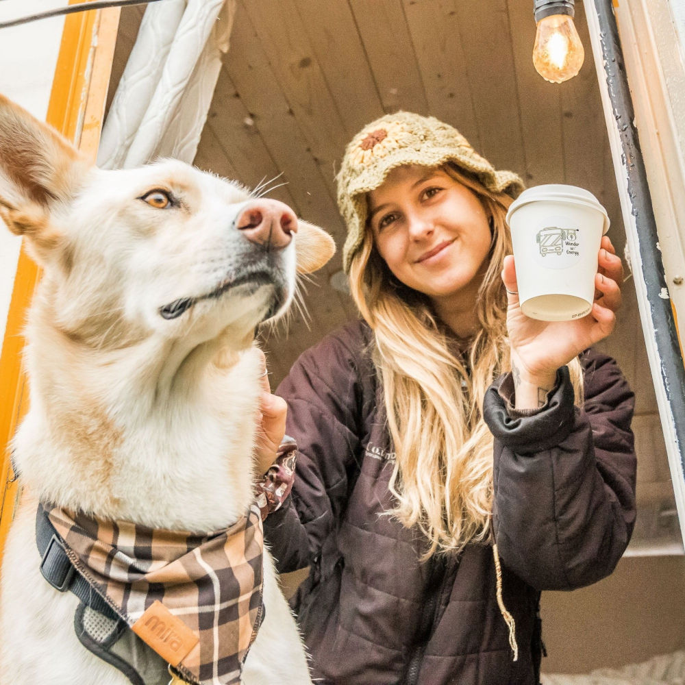 Woman in a knitted hat with a dog, both looking out the bus window, woman holding a Wonder with Energy cup.