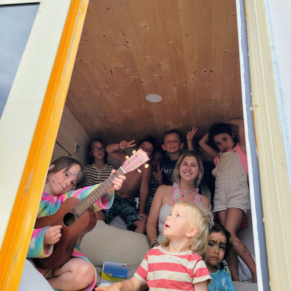 Group of children and a woman in the cozy, wood-paneled interior of the bus, one child playing a ukulele.